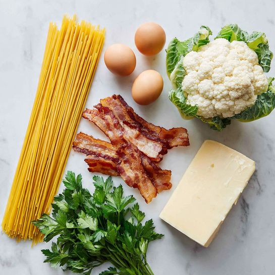 The image shows ingredients laid out on a white marbled surface. There is a bundle of uncooked spaghetti with a yellow color placed vertically on the left side. Next to it are three light brown eggs in a triangular arrangement. Toward the center right, a bunch of crispy bacon strips with a mix of brown and white fat is placed vertically. Above the bacon, there is a whole white cauliflower with green leaves around its base. To the right of the bacon, a large wedge of pale yellow cheese is positioned. In the bottom left corner, some bright green parsley leaves are spread. Photo taken with an iphone --ar 4:5 --v 7