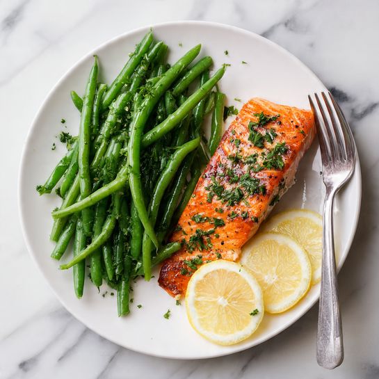 A white plate on a white marbled surface holds a dish with three main parts. On the left side, there is a pile of bright green beans with a fresh, smooth texture. Next to the green beans on the right, there is a cooked salmon fillet with a deep orange color and a slightly crispy, dark brown top layer sprinkled with small green parsley leaves. One lemon slice garnishes the top right of the salmon, and another lemon slice sits next to the green beans, both showing a pale yellow color with visible juicy segments. A silver fork rests on the plate beside the salmon. Photo taken with an iphone --ar 4:5 --v 7