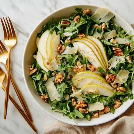 The image shows a white bowl filled with fresh green arugula leaves as the base layer, spread evenly inside the bowl. On top, there are two groups of thinly sliced pear pieces, placed neatly in a fanned-out pattern on opposite sides of the bowl. The pear slices are pale yellow with a slight green tint near the edges, and they have a glossy texture due to a light drizzle of dressing. The entire salad is sprinkled with coarse black pepper and small red pepper flakes, adding specks of dark and red colors over the green and pale yellow layers. The bowl sits on a white marbled surface. Photo taken with an iphone --ar 4:5 --v 7