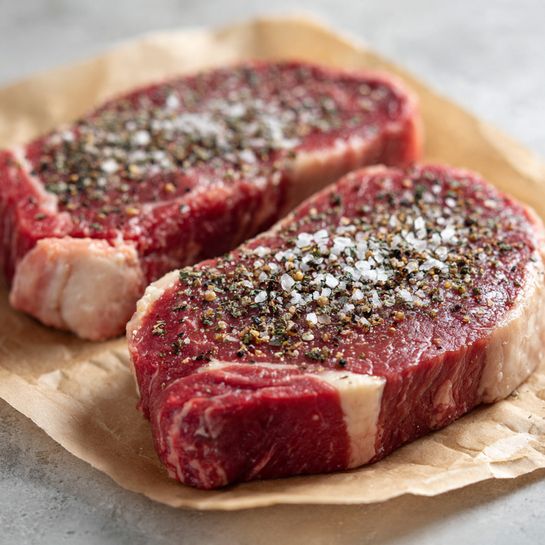 Two thick raw steaks are placed on light brown parchment paper with a white marbled surface underneath. Each steak is coated on top with coarse salt and cracked black pepper, showing a rich mix of red meat and creamy white fat along the edges. The texture of the meat is fresh and slightly marbled, with one steak positioned closer and the other behind it, both laying flat. Light softly illuminates the scene, creating a natural look. Photo taken with an iphone --ar 4:5 --v 7