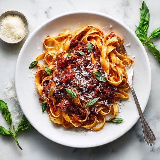 The image shows a white pan with a golden handle filled with pasta mixed with a chunky dark red sauce. The pasta is flat and light yellow, twisted in loops across the pan. The sauce has small pieces of red and dark brown vegetables that look soft and mixed evenly with the pasta. The inside sides of the pan have sauce stains, showing it has been cooked and stirred well. The pan sits on a white marbled surface with a small glass jar of grated cheese nearby, and a wine cork is placed above the pan. A soft white cloth with pink stripes is partly visible on the left side. The scene looks warm and fresh, with steam rising slightly from the dish photo taken with an iphone --ar 4:5 --v 7