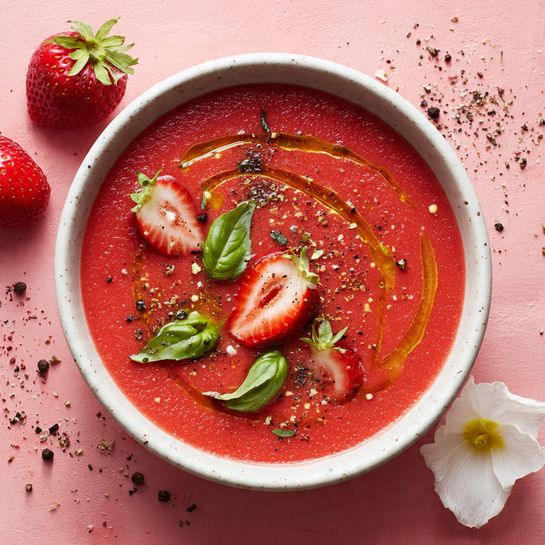 The image shows several white bowls filled with a smooth, deep red soup. Each bowl has two or three green basil leaves placed on the surface along with a light drizzle of golden oil forming a spiral pattern. The soup has tiny bubbles scattered on top, creating a fresh texture. Around the bowls, there are whole and halved fresh strawberries along with white flowers with light green centers, all arranged on a light pink surface. The scene is bright and colorful with a clean and fresh look. photo taken with an iphone --ar 4:5 --v 7