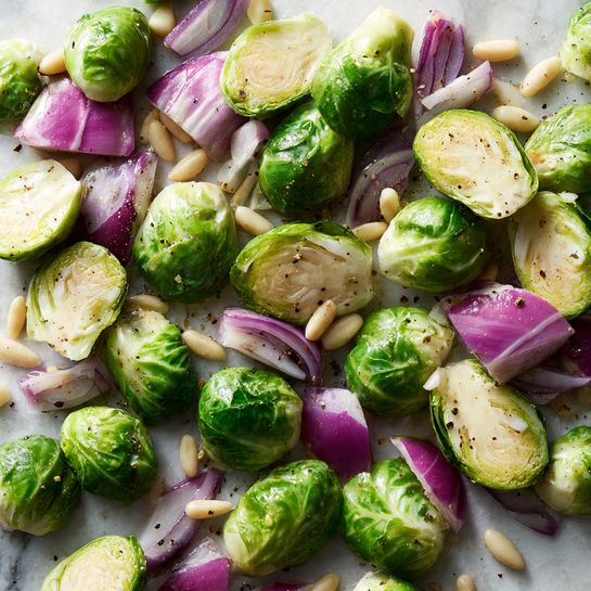 This image shows pieces of green Brussels sprouts cut into halves and quarters mixed with purple and white chopped onions scattered over a textured surface. Small white pine nuts are spread evenly among the vegetables. The Brussels sprouts have a fresh, crisp look with a light shine, and the onions are firm with layered sections visible. Some black pepper is sprinkled over everything, creating small dark specks, and there's a slight oily sheen covering the vegetables and pine nuts. The background is a white marbled texture. photo taken with an iphone --ar 4:5 --v 7
