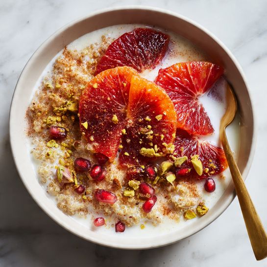 A white bowl filled with three main layers: at the bottom, cooked creamy quinoa mixed with small nuts and seeds, topped with bright red pomegranate seeds and pieces of chopped red fruit, all soaked in white milk creating a soft texture around the grains and fruits, and a gold spoon scooping up the mix. The bowl sits on a white marbled surface, and there is a white cup filled with light brown liquid in the background. Photo taken with an iphone --ar 4:5 --v 7