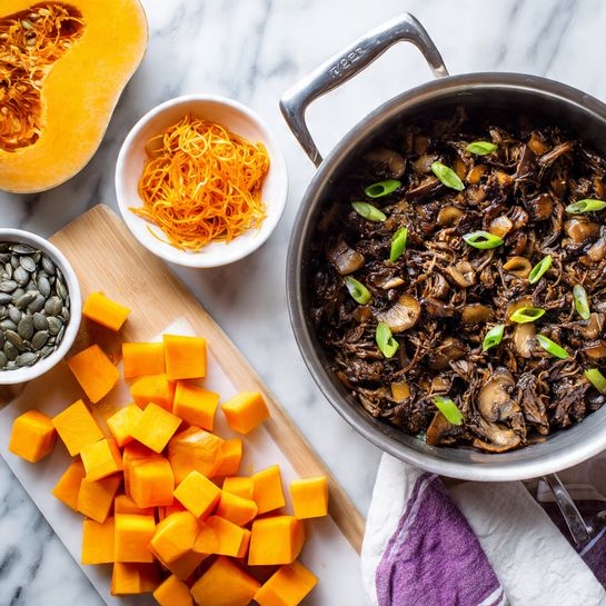A round silver pot filled with cooked shredded dark brown meat mixed with sliced mushrooms and small green onions is placed on a white marbled surface. Next to the pot, there is a white bowl with orange pumpkin seeds and orange pumpkin threads. In front, a white cutting board holds a half butternut squash with seeds inside and dozens of bright orange cubed squash pieces spread around it. A striped white and purple cloth is seen near the pot handle. Photo taken with an iphone --ar 4:5 --v 7