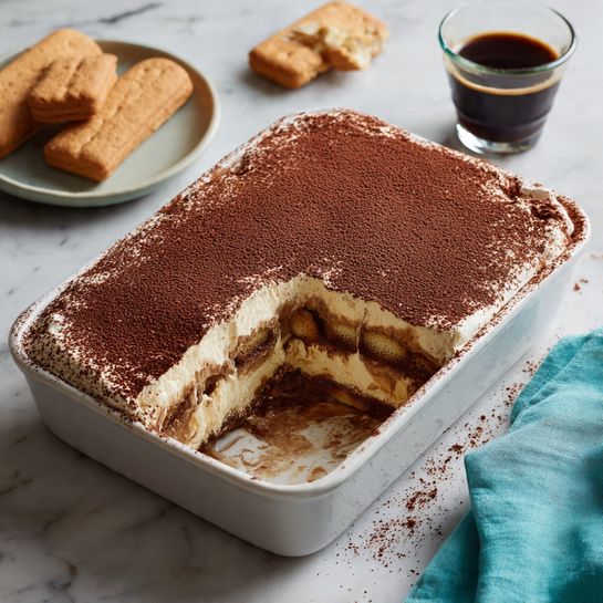 A rectangular white baking dish holds a layered dessert with one corner missing a square piece, showing three thick layers inside: a light cream on top, a darker brown layer in the middle, and another creamy layer below, all dusted evenly with dark cocoa powder. The surface of the dessert is smooth with a rich chocolate-brown color. The dish is placed on a white marbled surface with a few ladyfinger biscuits nearby, a small glass cup filled with dark coffee, and a teal cloth on the side. Photo taken with an iphone --ar 4:5 --v 7