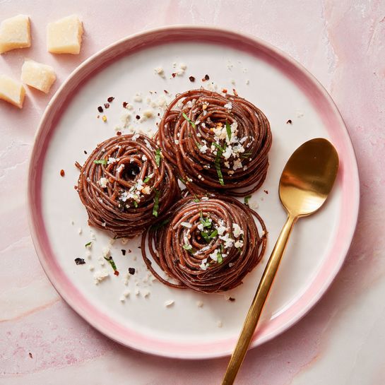 The image shows tightly coiled dark purple pasta nests placed closely on a white plate with a thin gold rim, lying on a white marbled surface. The pasta is topped with finely grated white cheese, small green herb pieces, and scattered red chili flakes, adding texture and color contrast. A gold fork rests beside the pasta on the plate, enhancing the elegant presentation. The dish has a rich, textured look with the glossy pasta spirals standing out against the smooth plate. Photo taken with an iphone --ar 4:5 --v 7