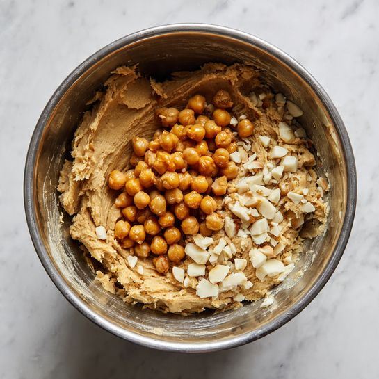 A silver metal mixing bowl sits on a white marbled surface, filled with a thick, light brown dough that takes up about half the bowl's space. On top of the dough, there are clusters of small round, orange-brown chickpeas grouped in the center, and chopped pieces of white macadamia nuts scattered to one side. The textures contrast between the smooth dough, shiny chickpeas, and rough chopped nuts. photo taken with an iphone --ar 4:5 --v 7