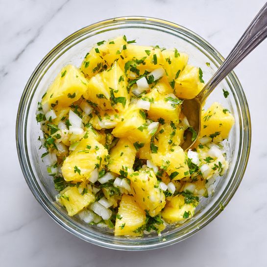 A clear glass bowl filled with a mix of bright yellow pineapple chunks, small pieces of white onion, and green chopped herbs, likely cilantro, giving it a fresh and colorful look. The pineapple pieces are the main layer, large and juicy, with onion bits and herbs spread evenly throughout, all sitting in a light liquid. A silver spoon is partially visible inside the bowl, slightly covered by the mixture. The bowl is placed on a white marbled surface. photo taken with an iphone --ar 4:5 --v 7