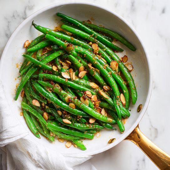 A white pan with a golden handle holds a dish of vibrant green beans scattered evenly across the pan. Mixed among the beans are golden brown toasted almond slices, adding texture and contrast. The green beans have a shiny, slightly oily surface, with small bits of seasoning visible. The pan rests on a white marbled surface, and a white cloth is wrapped around the handle, partially seen at the bottom of the image. photo taken with an iphone --ar 4:5 --v 7