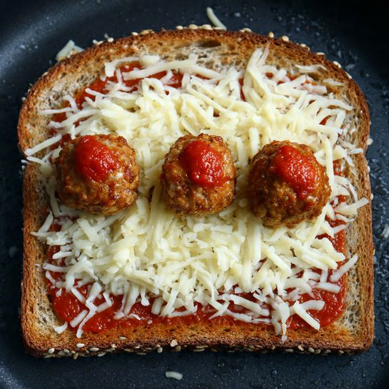 A single slice of multigrain bread with visible seeds on the crust sits flat in a dark pan. On top of the bread, there is a thick layer of white shredded cheese evenly spread. Above the cheese, three small brown meatballs are placed in a line, each topped with a small amount of bright red tomato sauce. The edges of the bread appear slightly soaked with oil or butter. The entire scene is set against a dark pan surface. Photo taken with an iphone --ar 4:5 --v 7