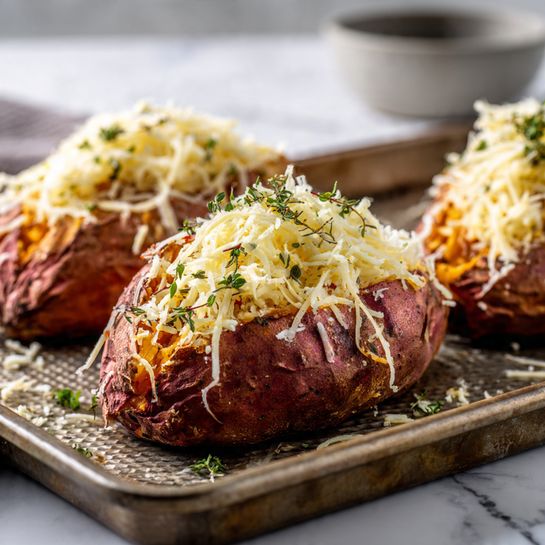 A close-up of three roasted sweet potatoes on a textured baking tray, each topped with a generous pile of shredded pale yellow cheese, some of which is falling onto the tray. The sweet potatoes have a rough, cracked, reddish-brown skin with soft spots showing, and the shredded cheese has a fine, stringy texture with small green herb flakes mixed in. The tray rests on a white marbled surface that is softly blurred in the background. Photo taken with an iphone --ar 4:5 --v 7
