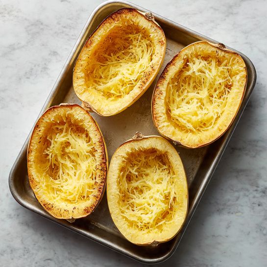 The image shows four halves of a cooked spaghetti squash placed inside a metal baking tray. Each half displays a rough, stringy texture with long, pale yellow fibers inside the hollowed center. The outer squash skin is a soft yellow color with a slightly browned edge from cooking. The baking tray rests on a white marbled surface that adds subtle light reflections around it. photo taken with an iphone --ar 4:5 --v 7