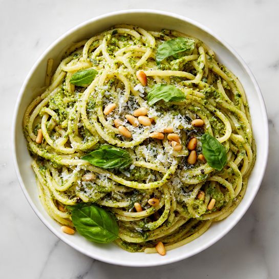 A close-up view of a large white bowl filled with spaghetti covered in green pesto sauce. The spaghetti strands are twisted and layered, with an uneven spread of bright green pesto and small toasted pine nuts scattered on top. Fresh basil leaves add a pop of vibrant green color across the surface, and grated parmesan cheese is lightly sprinkled over the dish, creating a fine white texture. The bowl sits on a white marbled surface, and the overall look is fresh and appetizing. Photo taken with an iphone --ar 4:5 --v 7
