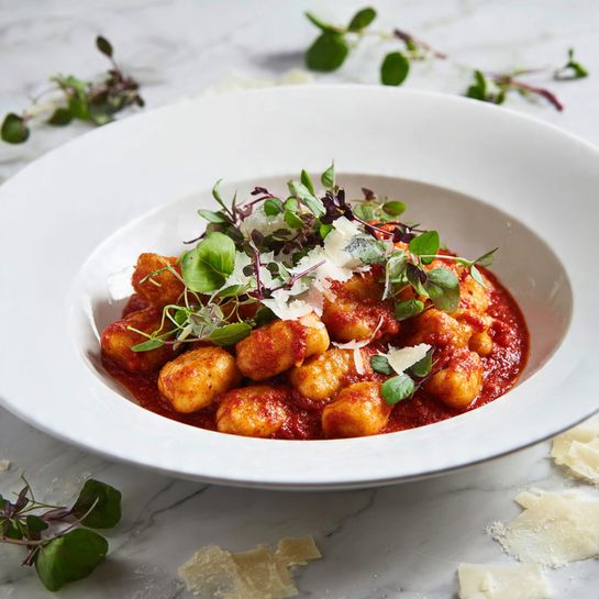 A white bowl filled with golden brown gnocchi pieces that have a slightly crisp texture on the outside, all coated in a rich red tomato sauce. The gnocchi sit in a shallow layer of the sauce, which has a smooth appearance. Scattered on top are small green leaves of fresh herbs and thin, irregular white cheese shavings that add texture and contrast to the warm colors of the dish. The bowl is placed on a white marbled surface with some cheese shavings and herb leaves around it. Photo taken with an iphone --ar 4:5 --v 7