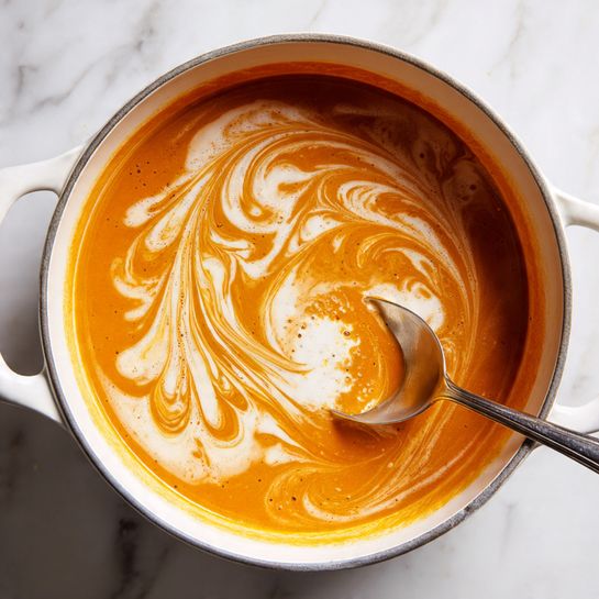 A close-up of a white pot filled with a thick orange soup or sauce being stirred by a silver spoon. The soup has swirling patterns of creamy white foam on the surface, creating a marbled effect. The pot rests on a white marbled surface. Photo taken with an iphone --ar 4:5 --v 7