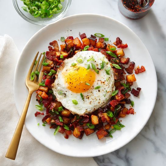 The dish shows a base layer of golden brown roasted vegetables with orange sweet potatoes and green zucchini pieces mixed with crispy pieces of browned bacon, all piled in the center of a white plate sitting on a white marbled surface. On top of the vegetable and bacon mix is a fried egg with a bright yellow runny yolk spilling over the layers beneath. Scattered small green onion slices and black pepper specks decorate the surface around and over the food, adding color and texture contrast. photo taken with an iphone --ar 4:5 --v 7
