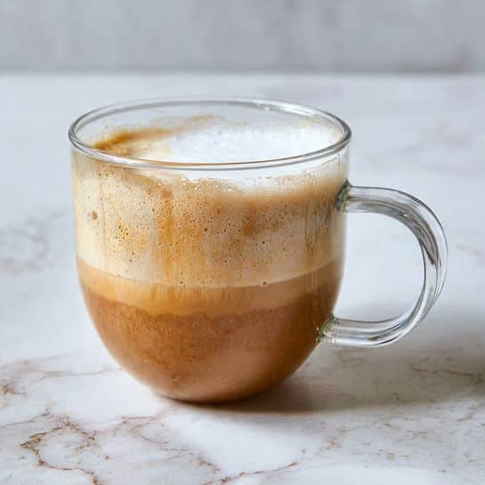 A clear glass cup with a handle holds two visible layers visible from above: one layer is light brown and smooth, covering most of the cup, and the other is thick white foam, located on the right side with a slightly glossy surface and soft texture. The glass cup rests on a white marbled surface, with a bit of textural pattern in the background. photo taken with an iphone --ar 4:5 --v 7