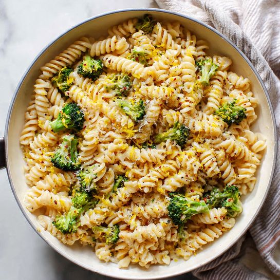 A close-up view of a white pan filled with spiral pasta that is creamy light beige in color, mixed with small green broccoli florets scattered evenly on top and throughout. The pasta has a smooth texture with slight shine, and is sprinkled with small bits of grated cheese and specks of black pepper. Bright yellow lemon zest is lightly spread over the dish, adding small pops of color. The pan rests on a white marbled surface with a soft striped cloth nearby. Photo taken with an iphone --ar 4:5 --v 7