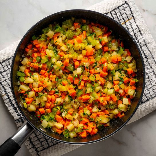 A black pot filled with mixed diced vegetables is shown. The vegetables include light yellow potatoes, bright orange carrots, and green celery, all chopped into small cubes. There are some small pieces of onion and green herbs scattered evenly throughout the mixture. The pot is placed on a folded white cloth with a black grid pattern, which sits on a white marbled surface. The image is bright and clear, highlighting the colors and textures of the fresh vegetables, photo taken with an iphone --ar 4:5 --v 7