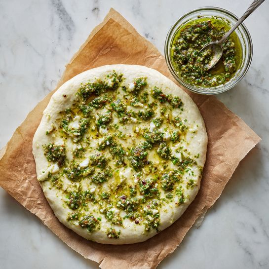Artichoke Burrata Pizza with Lemon Basil Pesto Recipe 4 A round, uncooked pizza dough sits flat on a piece of brown parchment paper, resting on a white marbled surface. The dough is pale and smooth, with patches of green pesto sauce spread unevenly on top, showing varied textures with small bits in the sauce. To the upper right of the dough, there is a small clear glass bowl filled with more green pesto, a silver spoon resting inside it with some pesto on the spoon. The whole scene is lit softly, highlighting the fresh dough and vibrant green pesto. photo taken with an iphone --ar 4:5 --v 7
