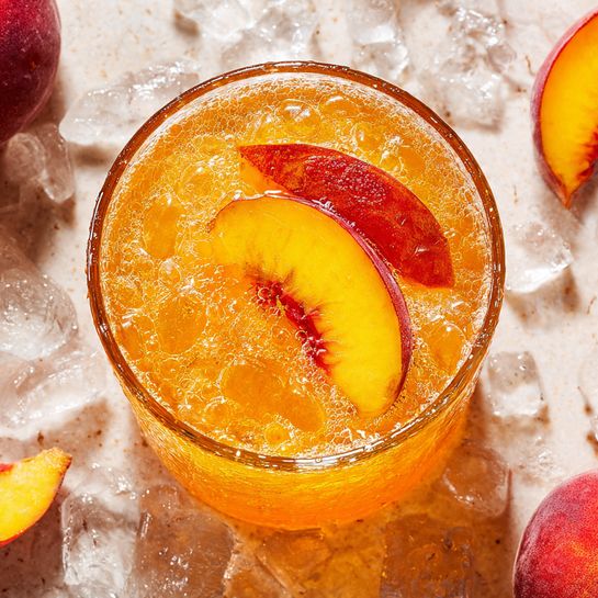 The image shows two glasses of a peach-colored drink on a white marbled surface scattered with ice pieces. The glass on the left is round and short, filled with ice and the orange drink, with some small peach pieces inside. The glass on the right is tall and wide at the top like a wine glass, also filled with ice and the same orange drink, containing a bigger slice of peach inside. Around the glasses are several peach slices and a few lemon wedges in the front, all resting on the white marbled surface. In the background, there is a whole peach and a blurred green outdoor setting. Photo taken with an iphone --ar 4:5 --v 7