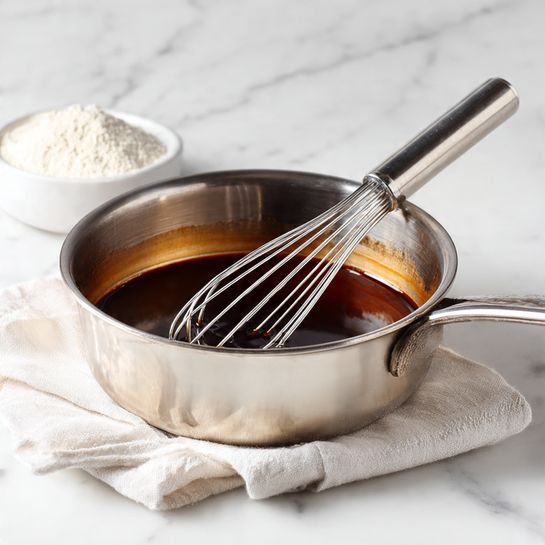 The image shows a shiny silver metal saucepan with dark brown liquid inside, which looks smooth and reflective. A silver whisk is resting in the pan, partially submerged in the liquid, with its handle leaning on the edge of the pan. The pan is placed on a white cloth, and in the background, there is a small white bowl with a white powdery substance. The whole setup is on a white marbled surface photo taken with an iphone --ar 4:5 --v 7