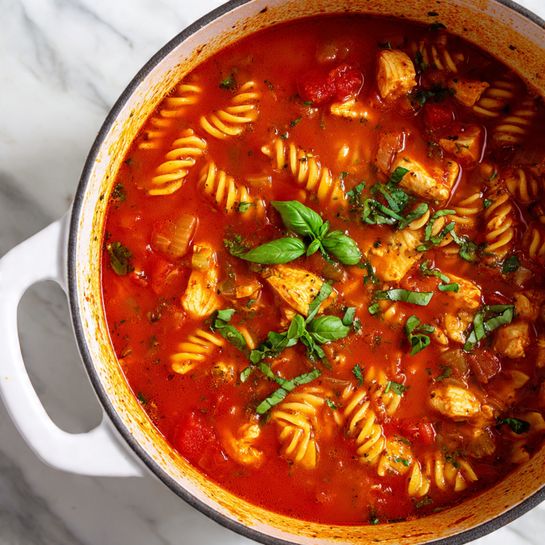 The image shows a white pot filled with a rich, red tomato soup that has spiral pasta and chunks of cooked chicken throughout. The soup has visible pieces of diced tomato and small onion bits, giving it a chunky texture. Fresh green basil leaves are placed on top, adding a pop of vibrant green color. The sides of the pot show sauce splatters, indicating the soup has been stirred well. The pot sits on a white marbled surface. photo taken with an iphone --ar 4:5 --v 7