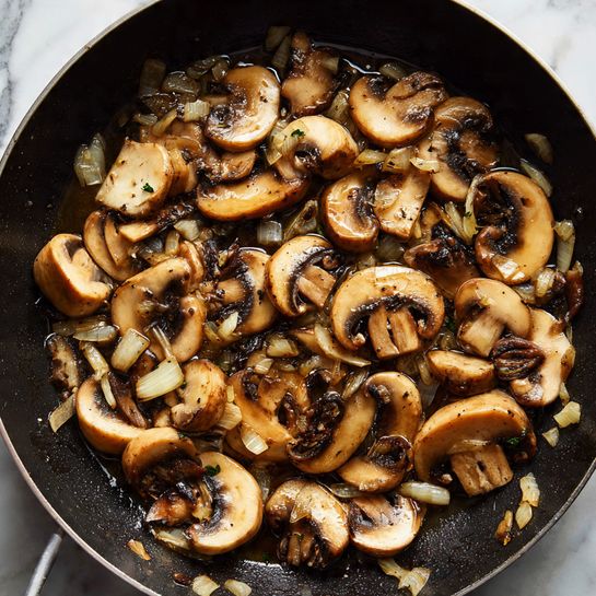 A close-up view inside a black pan filled with cooked, sliced mushrooms in liquid. The mushrooms are light brown and beige, with some pieces showing dark edges, mixed with small bits of translucent chopped onions scattered evenly throughout. The mushrooms have a soft, moist texture, sitting in a thin, shiny sauce that covers the bottom of the pan. The pan edges are slightly worn, and the focus is on the mix of mushrooms and onions inside. The background features a white marbled surface. photo taken with an iphone --ar 4:5 --v 7