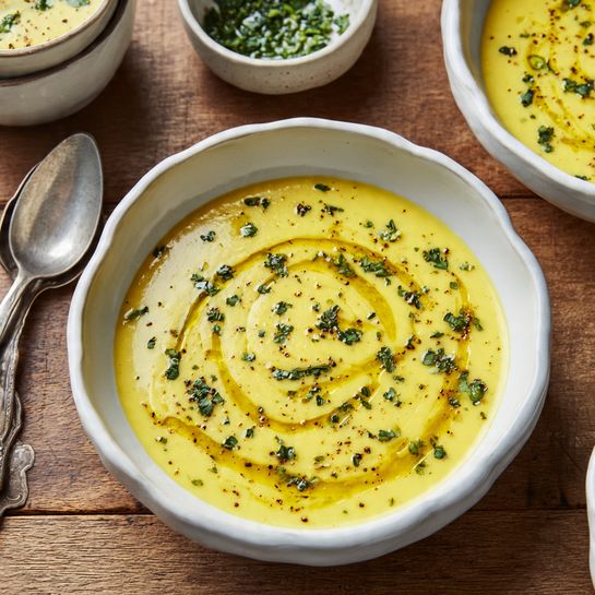 Four round small green pots filled with smooth yellow soup are placed on a white marbled surface. Each pot has two small handles, with the word