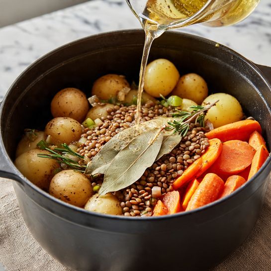 A close-up view of a large black pot filled with layered food. The bottom layer consists of small whole baby potatoes in light brown and yellow shades mixed with sliced orange carrots and bits of green vegetables. On top of this layer is a thick layer of dry light brown lentils, with two large light brown bay leaves placed on them. A light golden liquid is being poured into the pot from above. The pot sits on a white marbled surface, with a neutral beige cloth underneath. Photo taken with an iphone --ar 4:5 --v 7