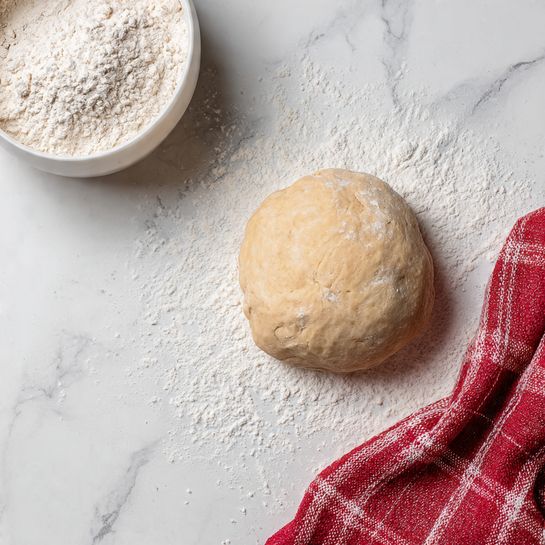 A round ball of light beige dough sits on a white marbled surface with white flour scattered lightly around it. To the top left, a white bowl filled with flour is partially in view. At the bottom right corner, there is a folded red and white checkered cloth. The scene is simple and bright, focusing on the dough's texture and the surrounding flour. Photo taken with an iphone --ar 4:5 --v 7