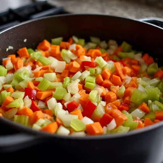 A close-up image showing a black pot filled with diced vegetables being cooked. The vegetables include bright orange carrots, light green celery, white onions, and red bell peppers. The pieces are small and roughly even in size, covering the bottom of the pot in a colorful mix. The pot sits on a stovetop, and the scene is lit well, showing the fresh colors and soft texture of the cooked vegetables. photo taken with an iphone --ar 4:5 --v 7