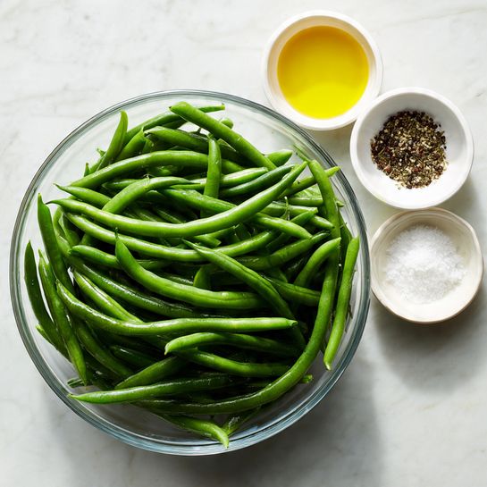 A clear glass bowl filled with fresh green beans, showing a rich green color and smooth texture. Above the bowl, there are three white small bowls arranged in a triangular shape; one contains a yellow liquid (olive oil), another has white salt, and the last holds black pepper. The entire setup rests on a white marbled surface with soft, natural lighting highlighting the freshness of the green beans and the simple ingredients. photo taken with an iphone --ar 4:5 --v 7