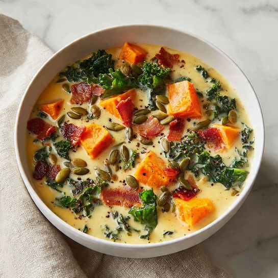This image shows a close-up of a creamy soup inside a white pot with a visible handle. The soup has three main layers: the creamy yellow broth that fills most of the pot, chunks of orange sweet potato or carrot scattered throughout, and dark green leafy kale pieces spread evenly on top and mixed in. The textures show the broth as smooth and slightly frothy, the orange chunks as soft yet solid, and the kale as wilted but still vibrant. The pot sits on a white marbled surface. photo taken with an iphone --ar 4:5 --v 7