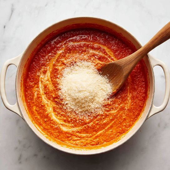The image shows a white pot filled with a thick, bright orange-red sauce with swirls of a lighter creamy color mixed in. In the middle of the sauce, there is a mound of fine white grated cheese sitting on the surface. A wooden spoon with a long handle rests inside the pot, partially dipped in the sauce and pointing towards the cheese. The pot is placed on a white marbled surface. photo taken with an iphone --ar 4:5 --v 7