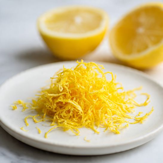 The image shows a close-up view of a small pile of bright yellow lemon zest placed at the center of a white plate with a smooth texture. Behind the zest, there is a lemon half with a pale yellow juicy interior, and part of the lemon peel is also visible to the side. The lemon zest looks finely shredded and slightly fluffy, with thin, stringy pieces that spread outward on the plate. The background and surface are a white marbled texture, slightly blurred to keep focus on the lemon zest and lemon half in front. Photo taken with an iphone --ar 4:5 --v 7