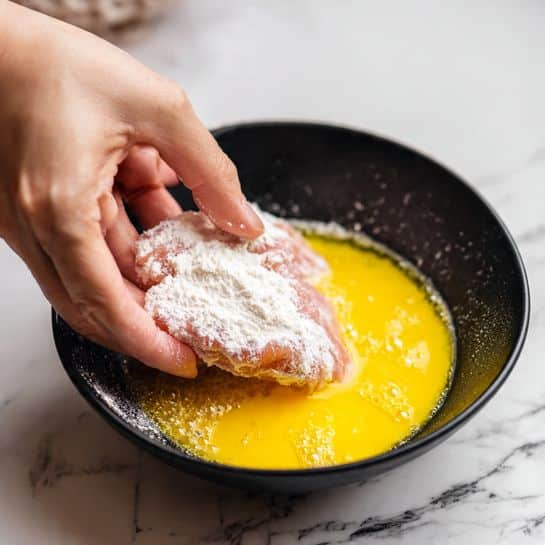 A close-up image showing a woman's hand dipping a piece of raw, pale pink meat coated in white flour into a shallow black bowl filled with bright yellow beaten eggs, with small bubbles on the egg surface. The coated meat piece covers most of the bowl's visible area. The background is a white marbled texture photo taken with an iphone --ar 4:5 --v 7