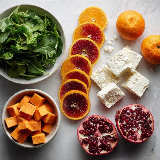 The image shows a flat lay of fresh ingredients on a white marbled surface. On the left side, there is a white bowl filled with leafy green salad. Next to it, a small white bowl holds cubed orange sweet potatoes. In the middle, there are slices of blood oranges arranged in a vertical line, showing bright red and orange colors in each slice. Beside the slices, there are chunks and a larger piece of white crumbly cheese scattered on the surface. On the right side, a pomegranate is cut in half, showing deep red seeds, with another piece of the fruit nearby. Whole oranges are placed around the composition for balance. The photo taken with an iphone --ar 4:5 --v 7