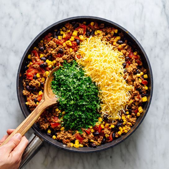 In a large black pan on a white marbled surface, there is a colorful mix of cooked ground meat, yellow corn, black beans, and small diced red tomatoes evenly spread around. On top of this mixture, on the right side, there is a fluffy pile of shredded yellow and white cheese, while on the left side, a bright green mound of chopped fresh herbs, likely cilantro, rests. A wooden spoon with visible wood grain is placed inside the pan to the left, partially touching the food, and a woman's hand is holding the pan's handle. The scene is bright and warm, showing the textures of the cooked ingredients clearly, photo taken with an iphone --ar 4:5 --v 7