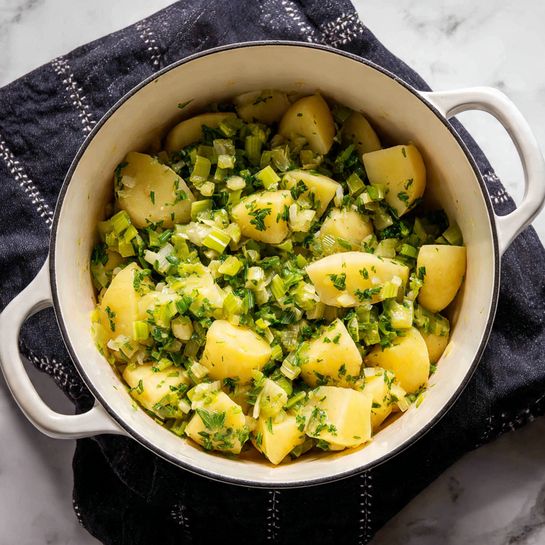 The image shows a large white pot filled with a mix of chopped yellow potatoes and green vegetables, including celery and onion. The potatoes are cut into medium-sized chunks with a smooth texture and pale yellow color. The green vegetables are chopped into smaller pieces, scattered evenly throughout the pot, adding a fresh, vibrant green contrast with various shades from light to medium green. The ingredients fill the pot almost to the top. The pot sits on a dark cloth with white stitching details, and the background is a white marbled surface. Photo taken with an iphone --ar 4:5 --v 7