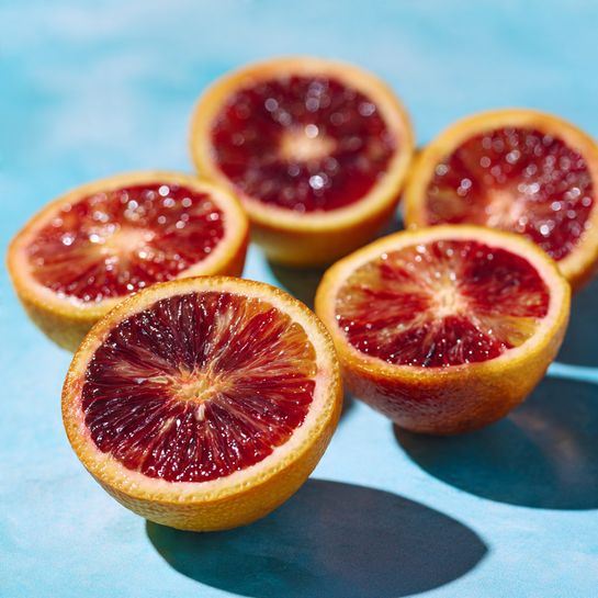 Six halves of blood oranges are spread out on a smooth, bright blue surface. The outer skin of the oranges is yellow with a slightly rough texture, while the inside flesh shows shades of deep red and lighter red with a juicy and segmented look. The halves are arranged in a loose group with some closer to the front and others fading softly into the background. The photo has a clear focus on the front orange half, with the rest softly blurred, showing natural light reflections on the orange surfaces. photo taken with an iphone --ar 4:5 --v 7
