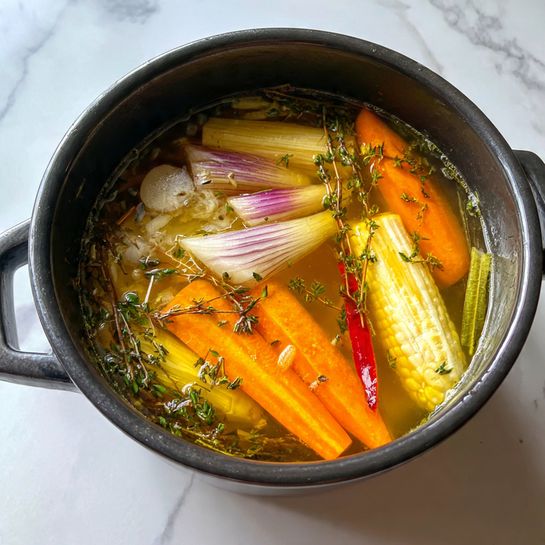 A black pot filled with clear yellow broth showing layers of soft white onion wedges on the top left, bright orange carrot sticks below, a piece of white corn on the cob on the upper right side, long thin red chili near the center, and scattered green leafy herbs and stems floating throughout. The pot sits on a white marbled surface. photo taken with an iphone --ar 4:5 --v 7