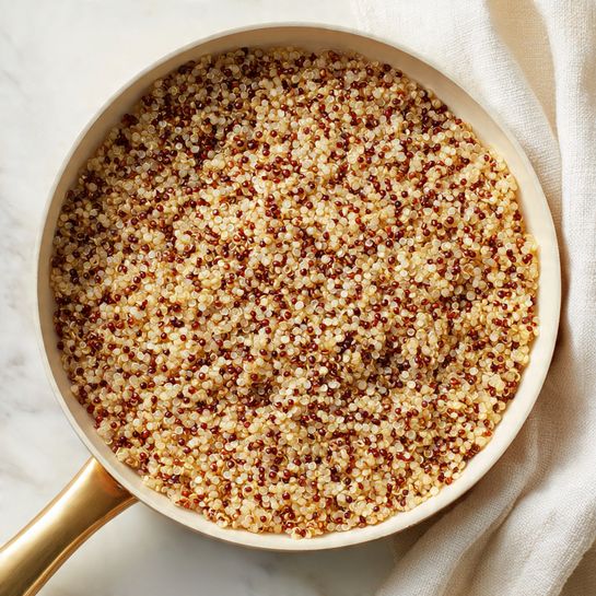 A close-up view of a white pan filled with cooked quinoa that has pale beige and dark red tiny round grains mixed together, creating a textured, grainy appearance. The quinoa covers the entire visible surface inside the pan, with slight clumps and variations in the distribution of colors. The pan has a shiny light gold handle that extends downward out of the frame. To the right bottom corner, there is a soft folded white cloth on a white marbled surface beneath the pan. The lighting brings out the fine details of each grain, giving a warm and natural look. Photo taken with an iphone --ar 4:5 --v 7
