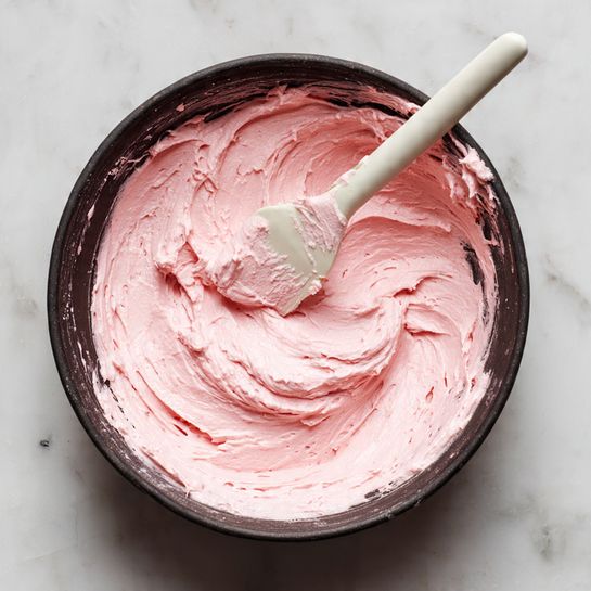 Inside a dark bowl, there is a smooth, thick, swirled mixture with a soft pink color. The creamy texture fills the center and spreads unevenly up the sides of the bowl, showing lighter and darker pink streaks. A white spatula with some pink mixture on it rests on one side, partially inside the bowl. The bowl is placed on a white marbled surface. photo taken with an iphone --ar 4:5 --v 7
