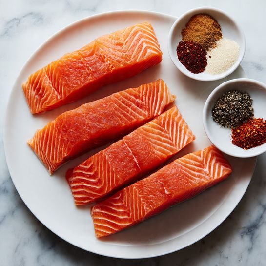 The image shows four bright orange pieces of raw salmon with visible fine white lines, placed on a white plate arranged closely together. Next to the plate on a white marbled surface, there is a small white bowl containing various spices in different colors: light beige, white, dark brown, black, and deep red. The spices are loosely grouped but remain separate from each other within the bowl. photo taken with an iphone --ar 4:5 --v 7