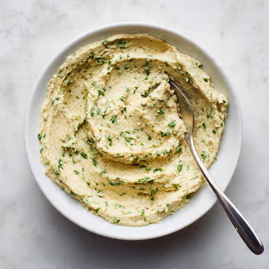 A shallow white bowl holds a single thick layer of a beige spread mixed with small green herb pieces throughout, creating a speckled texture. The spread covers the entire bowl evenly, with some swirled areas on top showing a creamy yet slightly coarse consistency. A shiny metal spoon rests inside the bowl, its round head partially covered with the spread, and the long handle extending out toward the bottom of the image. The bowl is placed on a white marbled surface. photo taken with an iphone --ar 4:5 --v 7