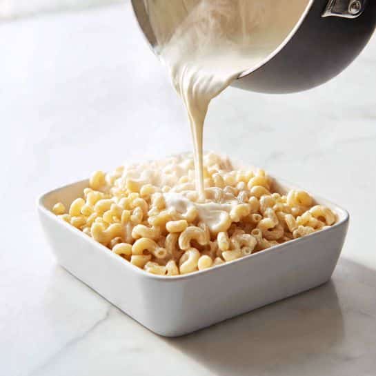 A black pot is tilted over a white square dish filled with cooked elbow macaroni pasta. White creamy sauce is being poured from the pot onto the golden-beige pasta, creating a smooth, thick stream covering the top layer of the pasta. Steam rises gently from the pot, adding a warm, fresh-cooked feeling. The dish sits on a white marbled surface with soft natural light shining on it. photo taken with an iphone --ar 4:5 --v 7
