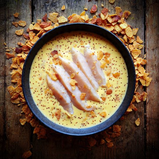 A dark bowl filled halfway with a light yellow creamy batter speckled with tiny brown dots, topped with several pale, smooth raw chicken strips resting in the middle. The bowl sits on a wooden surface scattered with corn flakes pieces. The overall look is simple and fresh, showing raw ingredients before cooking. Photo taken with an iphone --ar 4:5 --v 7