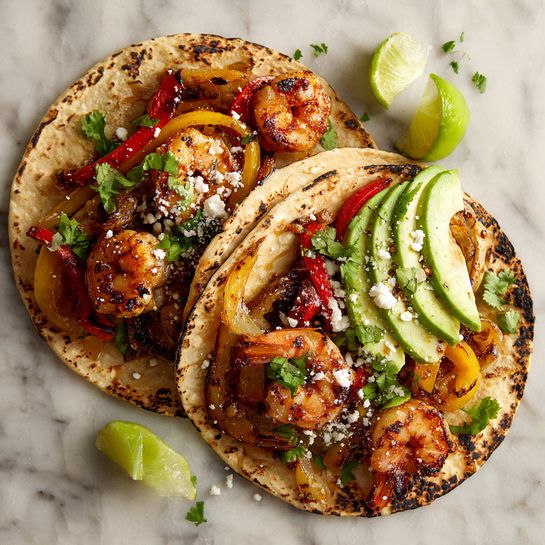 A single charred tortilla lies flat on a white marbled surface, topped with two grilled shrimp that are a warm orange and slightly shiny in texture. Beneath the shrimp, there are small strips of red and dark sautéed peppers mixed with bits of green cilantro leaves. White crumbly cheese is sprinkled over the shrimp and vegetables. To the right of the tortilla, a wedge of bright green lime rests partially on the edge. The background also shows some blurred lime slices and small scattered cilantro leaves. Photo taken with an iphone --ar 4:5 --v 7
